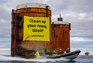 Greenpeace banner on oil structure in ocean, reads "Clean up your mess, Shell!", with Greenpeace activists in boat in foreground