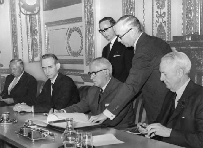 Men sitting and standing at a table, with one in centre signing a page