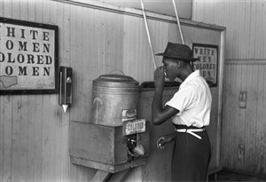 Man drinking at segregated water fountain with signs on the wall reading 'white women, colored women' and 'white men, colored men'